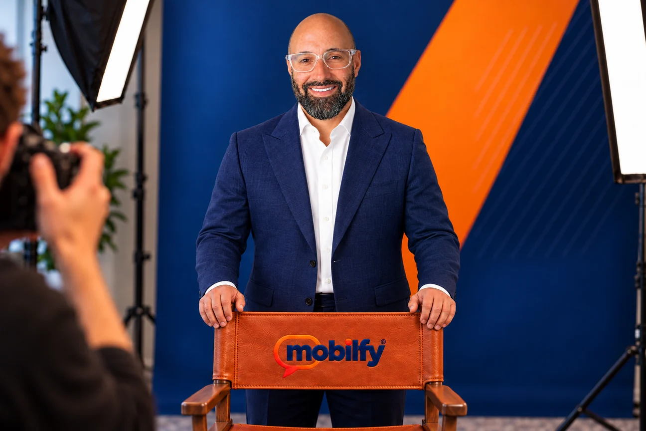 Angel Rivera in a studio portrait, standing behind a leather director’s chair embroidered with the Mobilfy logo