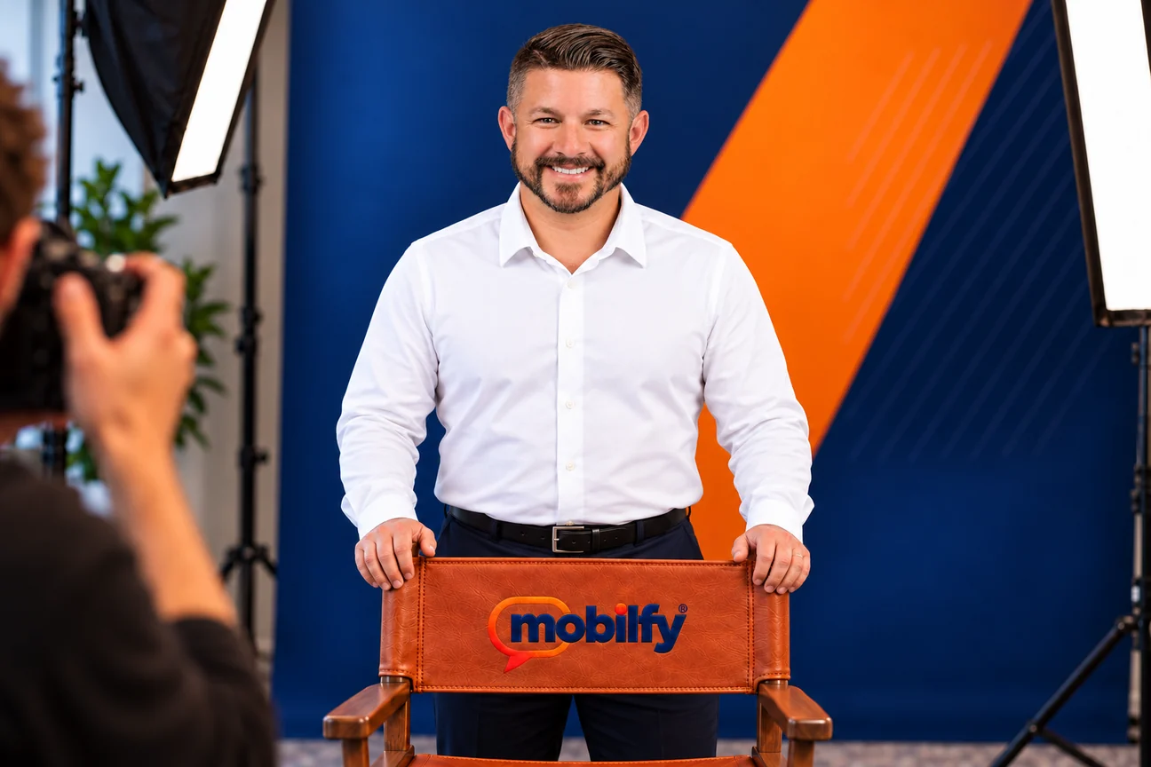 Will Powell in a studio portrait, standing behind a leather director’s chair embroidered with the Mobilfy logo