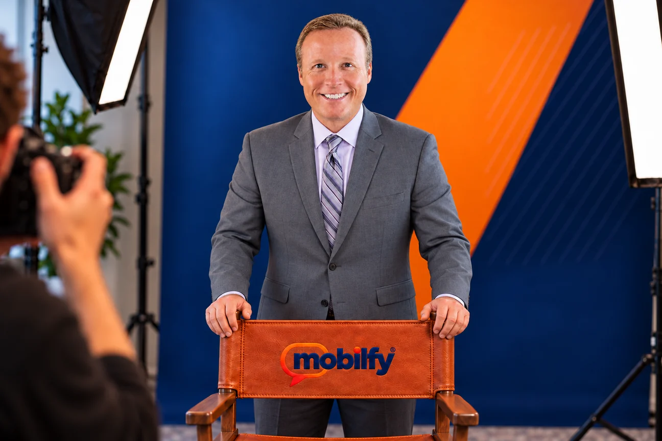William Davin in a studio portrait, standing behind a leather director’s chair embroidered with the Mobilfy logo